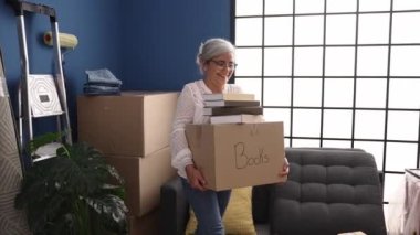 Middle age grey-haired woman smiling confident holding books package at new home