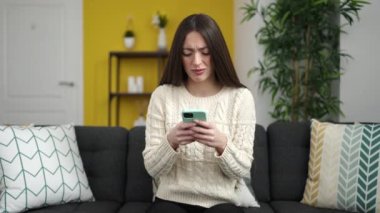 Young beautiful hispanic woman sitting on sofa with serious expression at home