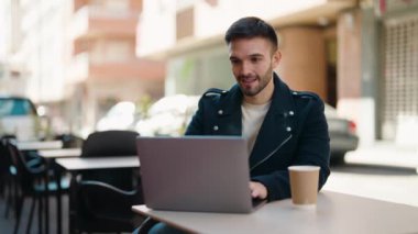 Young hispanic man using laptop sitting on table at coffee shop terrace