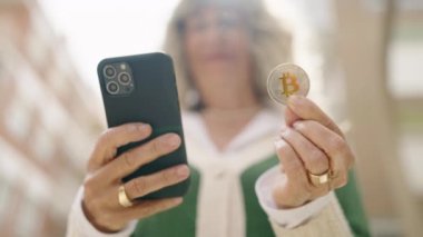 Orta yaşlı bir kadın sokakta Bitcoin tutarak akıllı telefon kullanıyor.