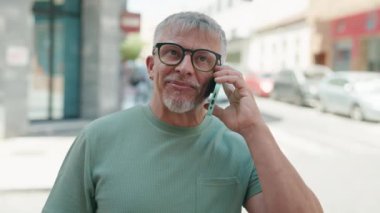 Orta yaşlı, kır saçlı, gülümseyen, kendine güvenen bir adam caddedeki akıllı telefondan konuşuyor.