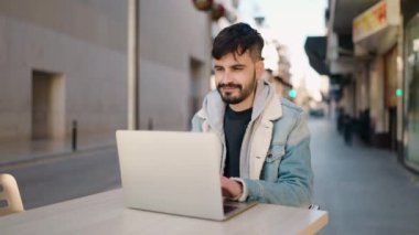 Young hispanic man using laptop sitting on table at coffee shop terrace