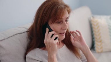 Middle age woman talking on the smartphone sitting on sofa at home