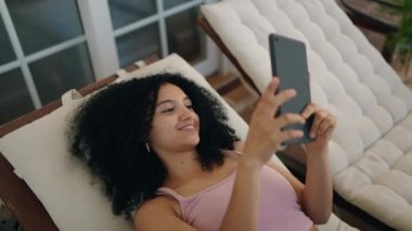 Young hispanic woman using touchpad lying on deck chair at home terrace