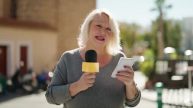 Middle age blonde woman reporter working using microphone reading notebook at street