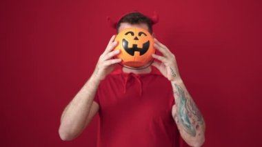 Young caucasian man wearing devil costume covering face with pumpkin over isolated red background