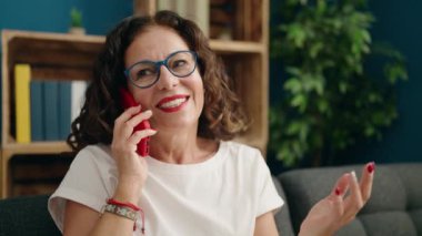 Orta yaşlı bir kadın akıllı telefonda konuşuyor. Evdeki kanepede oturuyor.