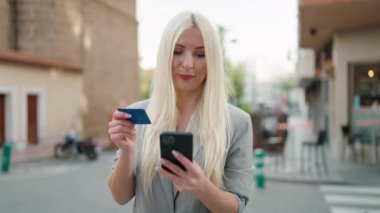 Young blonde woman executive using smartphone and credit card at street