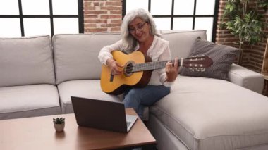 Middle age grey-haired woman smiling confident having online guitar class at home