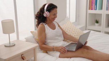 Middle age woman using laptop and headphones sitting on bed at bedroom