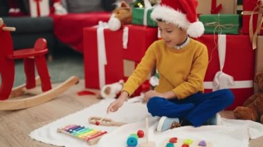 Adorable hispanic boy playing xylophone sitting on floor by christmas gifts at home