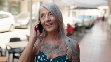 Orta yaşlı, kır saçlı, gülümseyen, kendine güvenen bir kadın caddedeki akıllı telefondan konuşuyor.