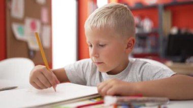 Adorable toddler student drawing on notebook sitting on table at classroom