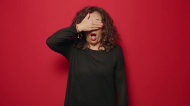 Middle age woman scared covering eyes with hand over red background