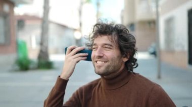 Young hispanic man listening voice message by smartphone sitting on wheelchair at street