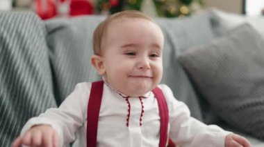Adorable hispanic baby sitting on sofa by christmas tree crying at home