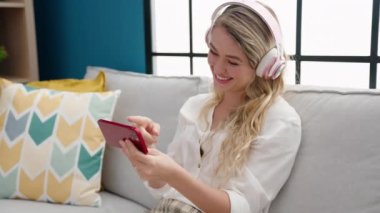 Young blonde woman watching video on smartphone sitting on sofa at home