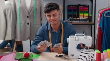 Young hispanic man tailor smiling confident holding dollars at clothing factory