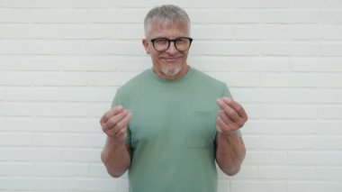Middle age grey-haired man smiling confident doing spend money gesture over isolated white brick background