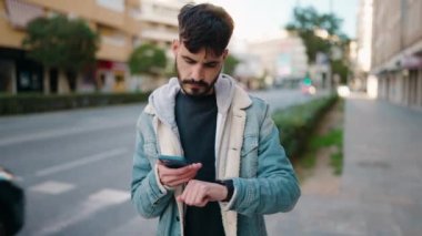 Young hispanic man using smartphone and looking watch at street