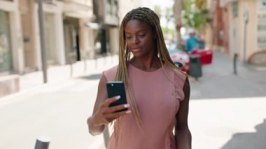Afro-Amerikalı kadın gülümsüyor. Sokakta akıllı telefonun yanında özçekim yapıyor.