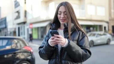 Genç, güzel, İspanyol bir kadın sokakta akıllı telefon kullanarak gülümsüyor.