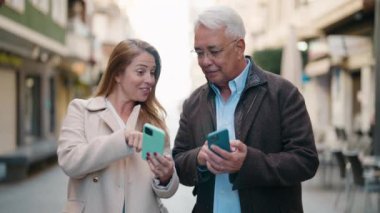 Middle age couple couple smiling confident using smartphone at street