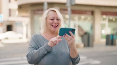 Middle age blonde woman smiling confident watching video on smartphone at street