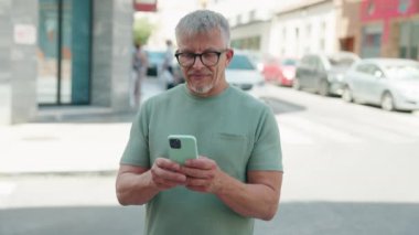 Orta yaşlı, kır saçlı, kendine güvenen, caddede akıllı telefon kullanan bir adam.