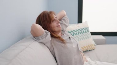 Orta yaşlı bir kadın, elleri başının üstünde, evinde kanepede oturuyor.