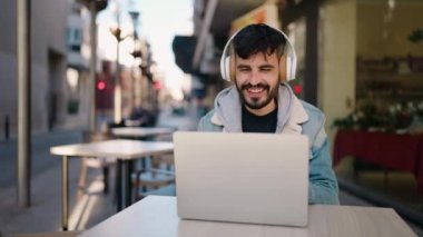 Young hispanic man listening to music sitting on table at coffee shop terrace