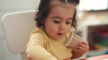 Adorable hispanic girl student sitting on table putting pencil color on cup at kindergarten