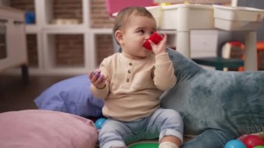Adorable toddler bitting plastic construction blocks sitting on floor at kindergarten