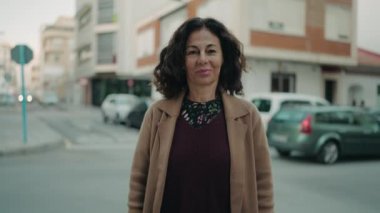 Orta yaşlı İspanyol kadın gülümsüyor. Kollarını kavuşturup caddede duruyor.