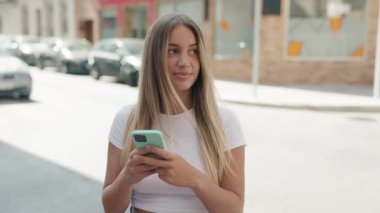 Genç, güzel, İspanyol bir kadın sokakta akıllı telefon kullanarak gülümsüyor.