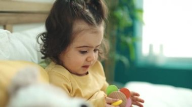 Adorable hispanic girl sucking toy sitting on bed at bedroom