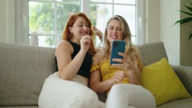 Two women using smartphone sitting on sofa at home
