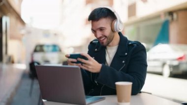 Young hispanic man playing video game sitting on table at coffee shop terrace