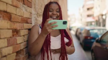 Afro-Amerikalı kadın gülümsüyor. Sokakta akıllı telefondan videolar izliyor.