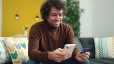 Genç İspanyol adam akıllı telefon ve kredi kartı kullanıyor. Evdeki kanepede oturuyor.