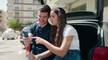 Young hispanic couple sitting on car trunk using smartphone at street