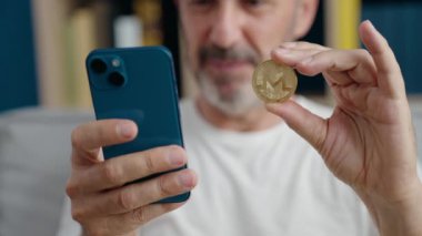 Monero Kripto 'yu elinde tutan orta yaşlı bir adam sokakta akıllı telefon kullanıyor.