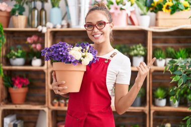 Young hispanic woman working at florist shop holding plant smiling happy pointing with hand and finger to the side 