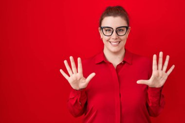 Young hispanic woman with red hair standing over red background showing and pointing up with fingers number ten while smiling confident and happy. 