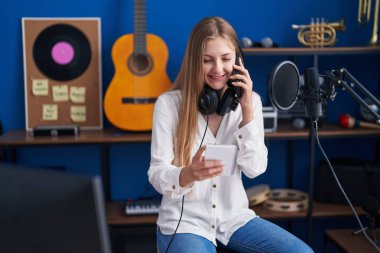 Young caucasian woman artist talking on smartphone reading notebook at music studio