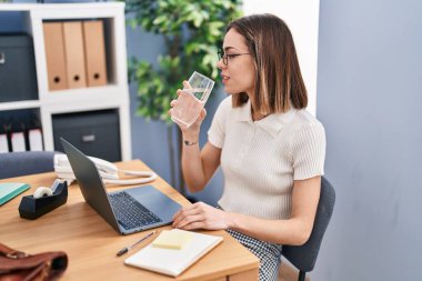 Young beautiful hispanic woman business worker using laptop drinking water at office