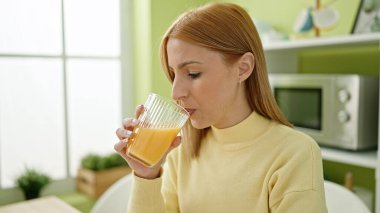 Young blonde woman drinking orange juice sitting on table at home