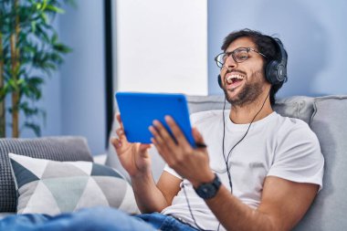 Young hispanic man watching video on touchpad sitting on sofa at home