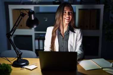 Young brunette woman working at the office at night with laptop looking away to side with smile on face, natural expression. laughing confident. 