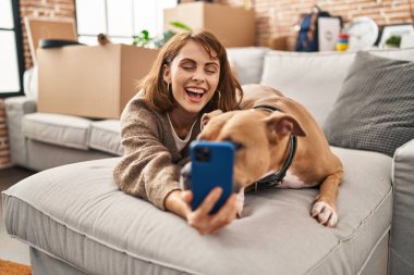 Kafkasyalı genç bir kadın yeni evinde köpekle kanepede uzanmış akıllı telefonuyla selfie çekiyor.